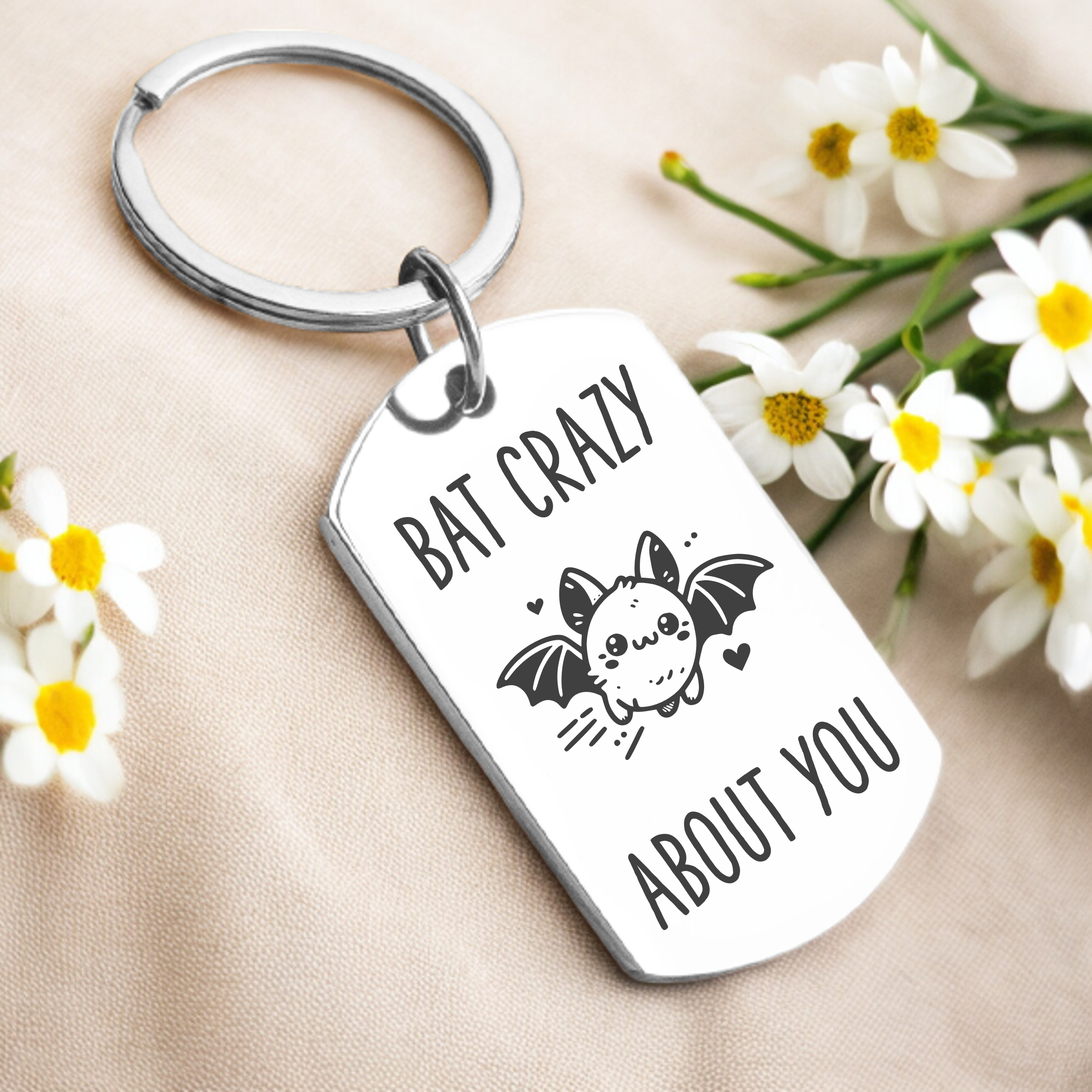 A personalized key ring with a silver or gold finish featuring a bat design and the phrase 'BAT CRAZY ABOUT YOU', presented alongside daisies.