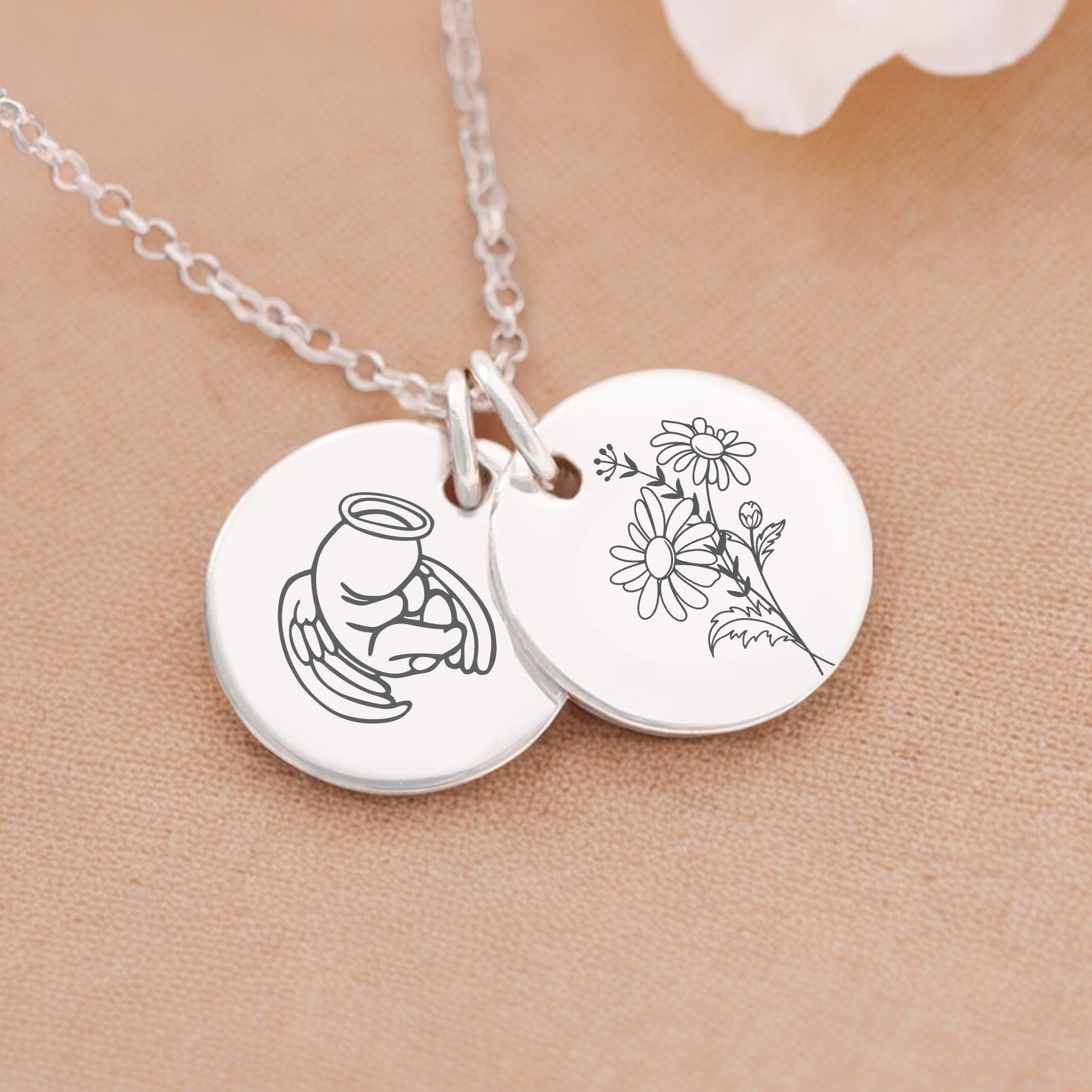 A silver necklace with two charms, one with an angel holding a flower and another with a floral design, displayed on a beige background with white flowers around it.