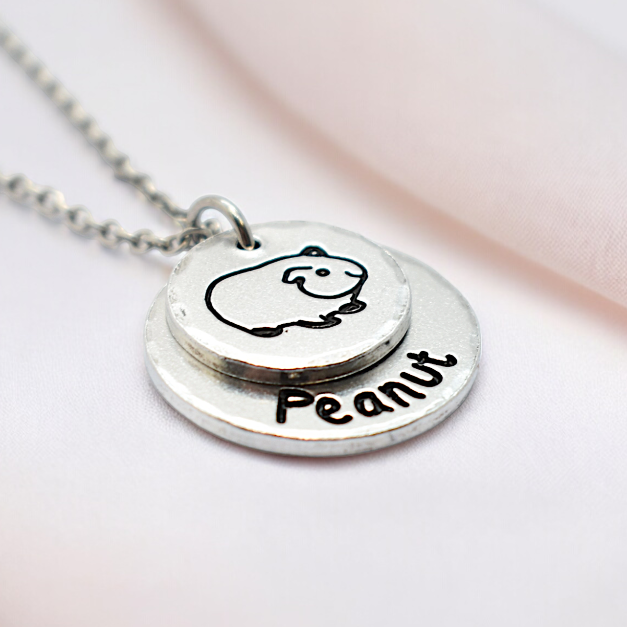A silver-colored necklace with a guinea pig design and the name 'Peanut' inscribed on a round tag, attached to a small chain.