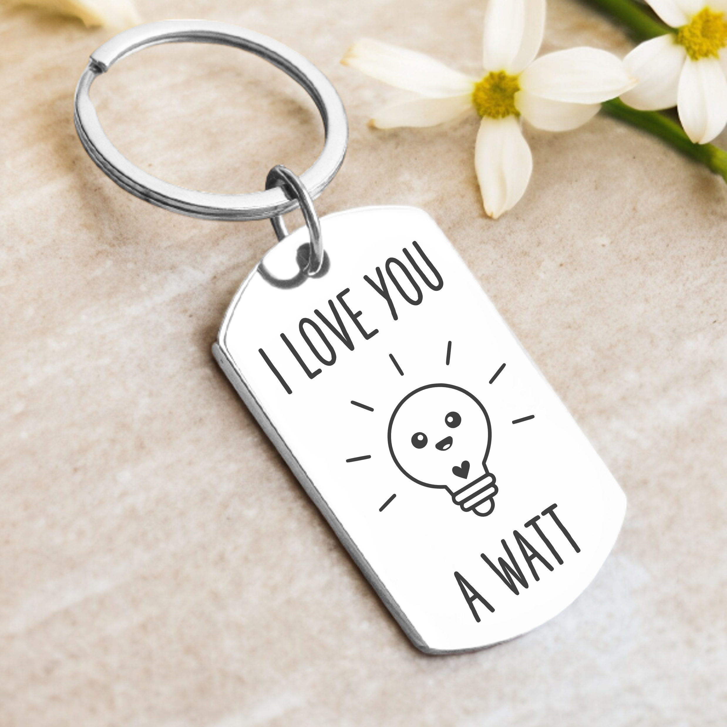 A key ring with a rectangular tag that has the phrase 'I LOVE YOU A WATT' printed on it, accompanied by a cartoon image of a light bulb. The tag appears to be made of silver and is displayed with a few petals at the bottom.