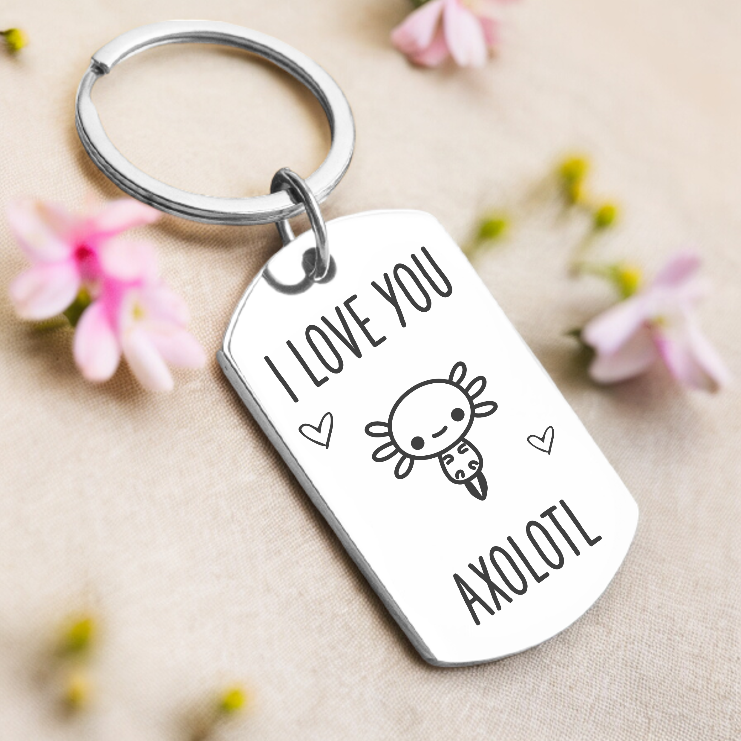 A key ring with a rectangular tag featuring an axolotl illustration and the text 'I love you axolotl'. The tag is attached to a key ring, and the background consists of a beige surface with some flowers scattered around.