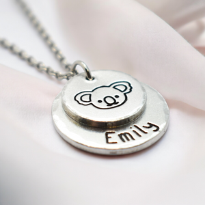 A silver-colored koala necklace with a personalized name 'Emily' stamped on it, presented against a pink background.