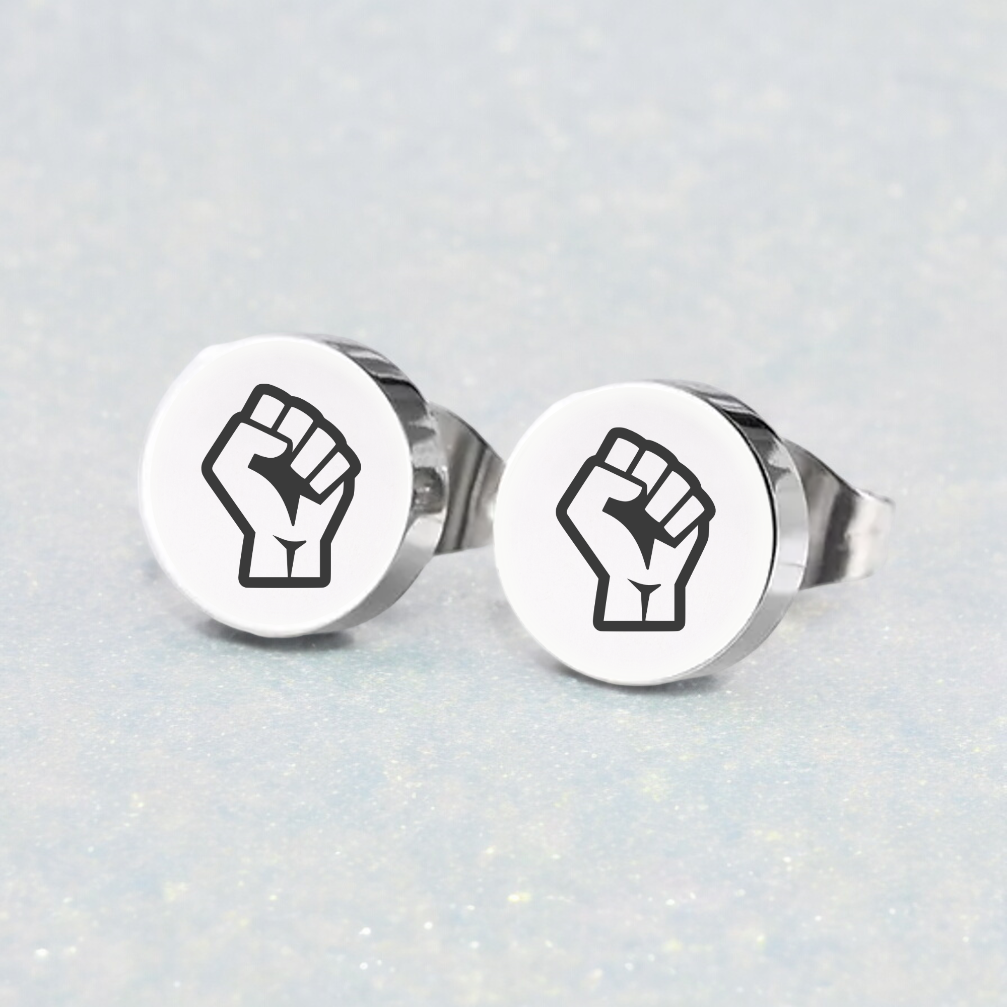 A pair of silver stud earrings featuring a black fist design on a white background, displayed against a sparkling background.