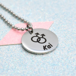 A silver-coloured pride necklace with a pendant featuring a black and white emblem with a male gender symbol and the name 'Kai' on it.