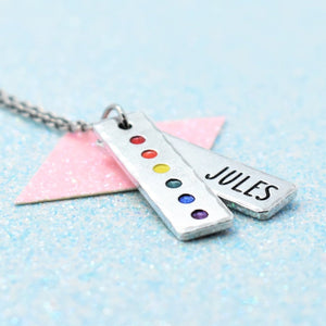 Customisable LGBTIQQ pride pendant with rainbow pride colours and 'JULES' engraved, on a light blue background.
