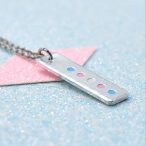 A minimalist pride necklace with a geometric shaped pendant featuring the transgender flag colours, attached to a silver chain.