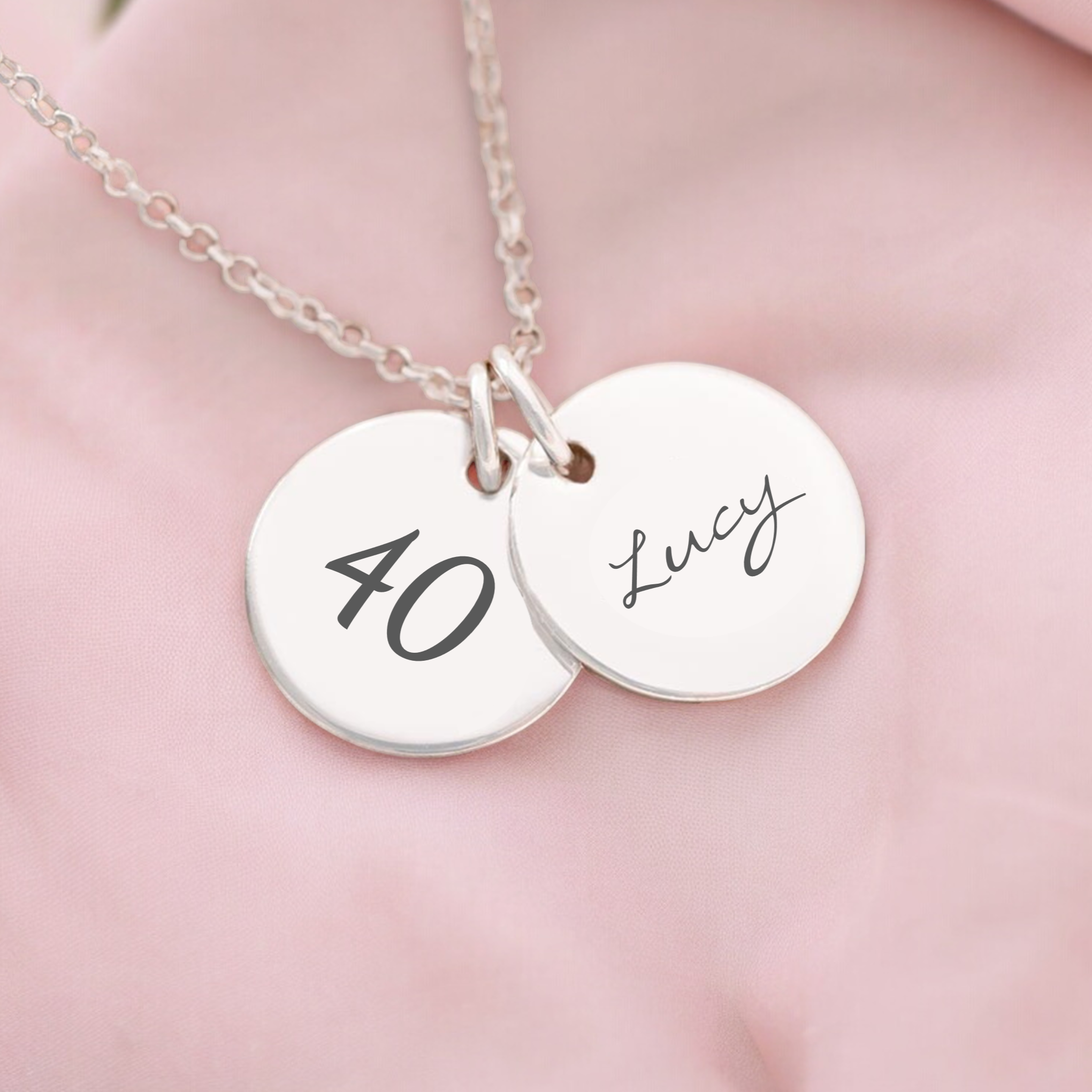 A personalized milestone necklace with two round pendants on a chain, one with the number 40 and the other with the name Lucy.