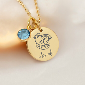 A gold birthstone necklace with a personalized engraving on a disc pendant, accompanied by a small blue gemstone, displayed on a white surface.