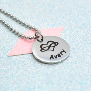 A polyamory necklace with a silver emblem and the name 'Avery' on it, attached to a pink tag.