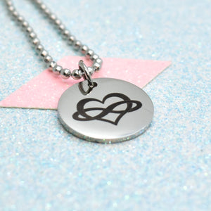 The image shows a silver-coloured necklace with a pendant featuring a black polyamory emblem. The pendant is round and has a shiny, metallic finish.