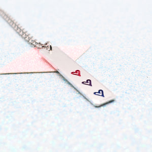 Necklace with a bar pendant on a ball chain featuring colourful hearts representing the bisexual pride flag on a light blue background