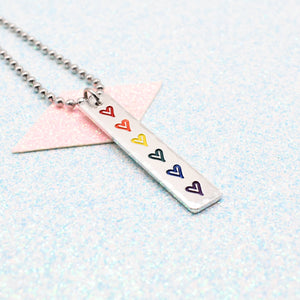 Necklace with a bar pendant on a ball chain featuring colourful hearts representing the traditional pride flag on a light blue background