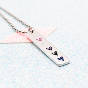 Necklace with a bar pendant on a ball chain featuring colourful hearts representing the gender fluid pride flag on a light blue background