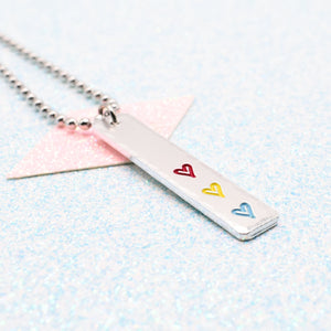 Necklace with a bar pendant on a ball chain featuring colourful hearts representing the pansexual pride flag on a light blue background