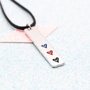 Necklace with a bar pendant on a faux leather cord featuring colourful hearts representing the polyamory pride flag on a light blue background