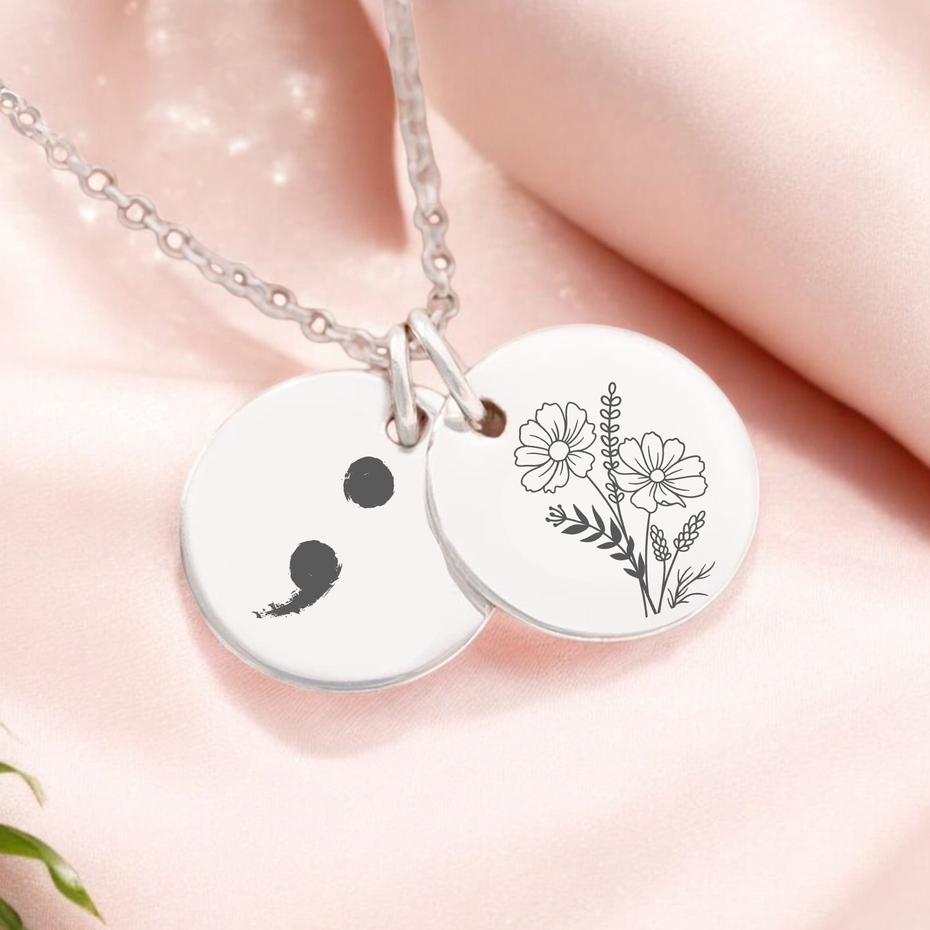 A necklace with two circular pendants, one featuring a semi-colon symbol and the other a birth flower, displayed on a pink background with some green leaves.