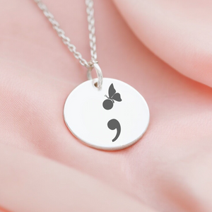 A semi-colon necklace with a butterfly charm on a person's finger, displayed against a pink background.