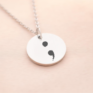 A semi-colon necklace with a silver pendant on a chain, displayed on a person's neck.