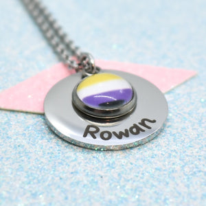 A custom pride necklace with a pride pendant featuring the non binary pride flag symbol, and a personalised name written on the pendant. The necklace is displayed on a blue background with a shimmering surface.
