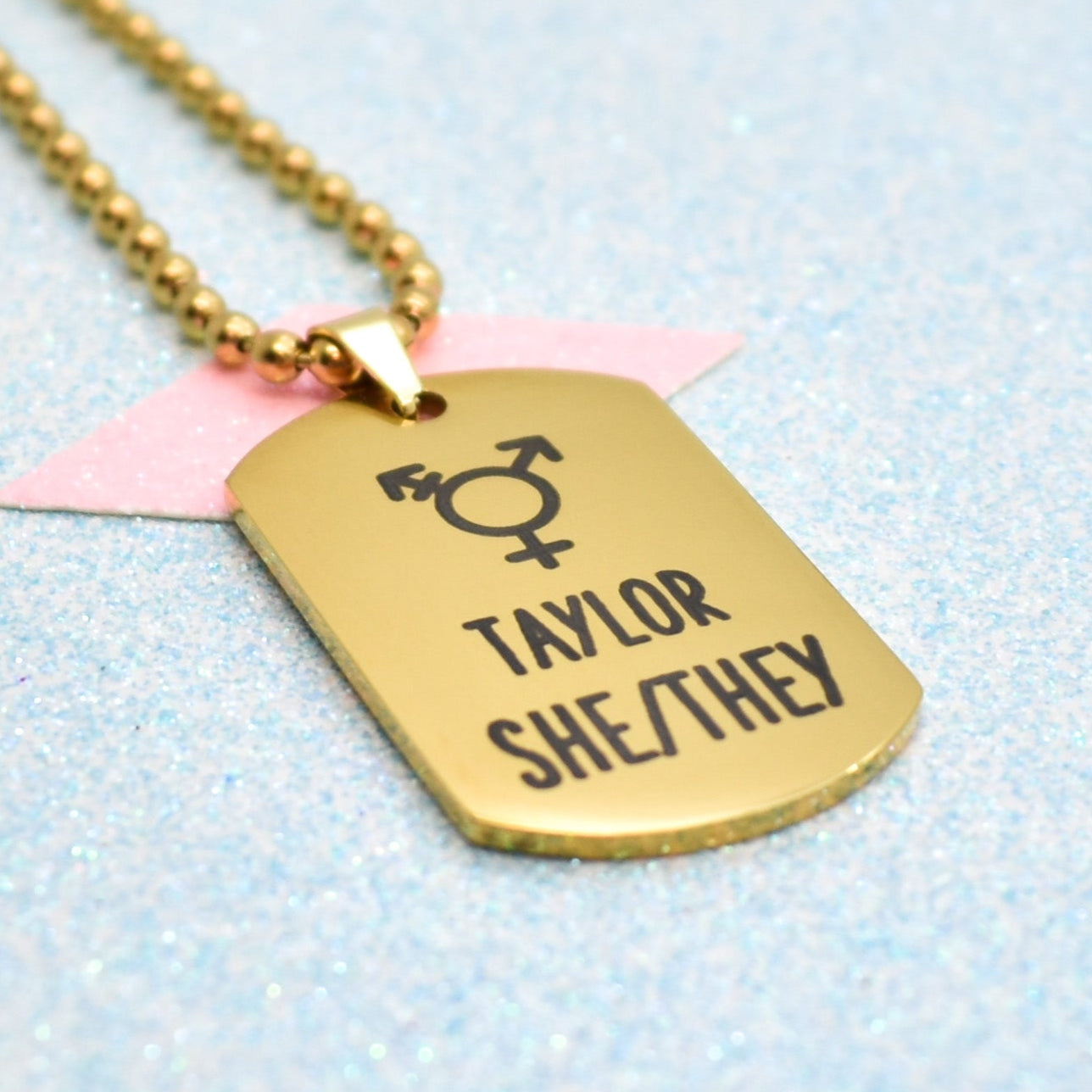 A gold-colored necklace with a pendant featuring the transgender symbol and the name 'Taylor Sheehy' on a sparkling blue background.