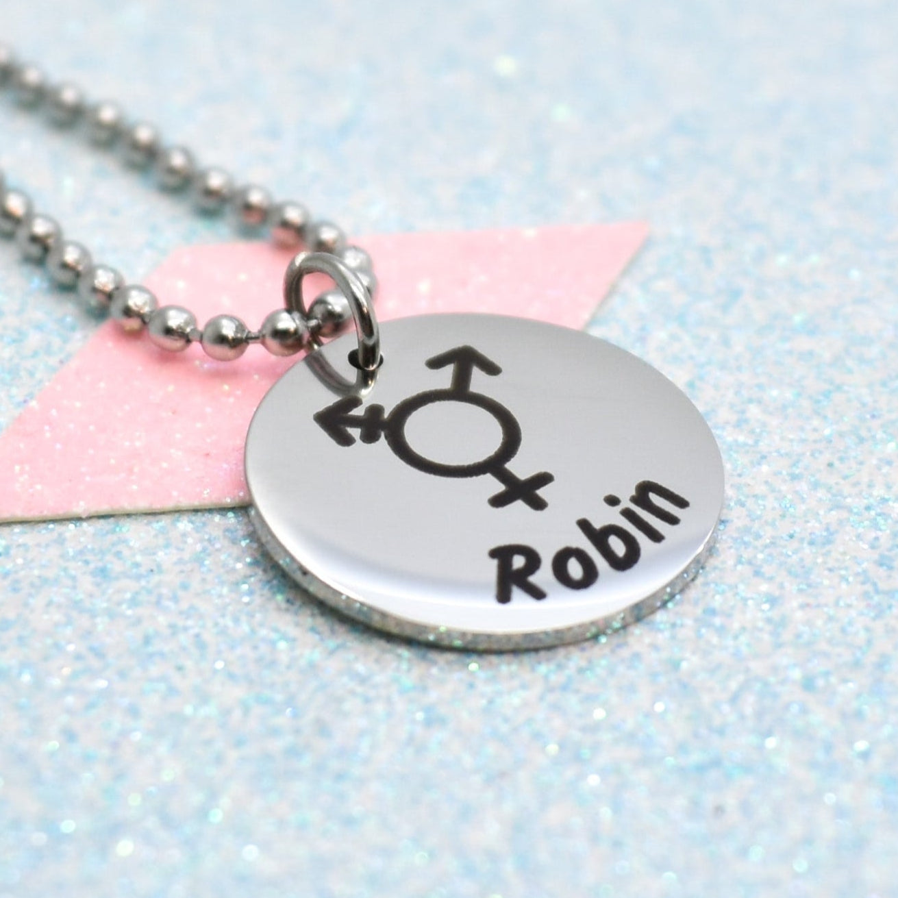 A transgender pride necklace with a silver emblem featuring a gender symbol and the name 'Robin' on it, attached to a silver chain.
