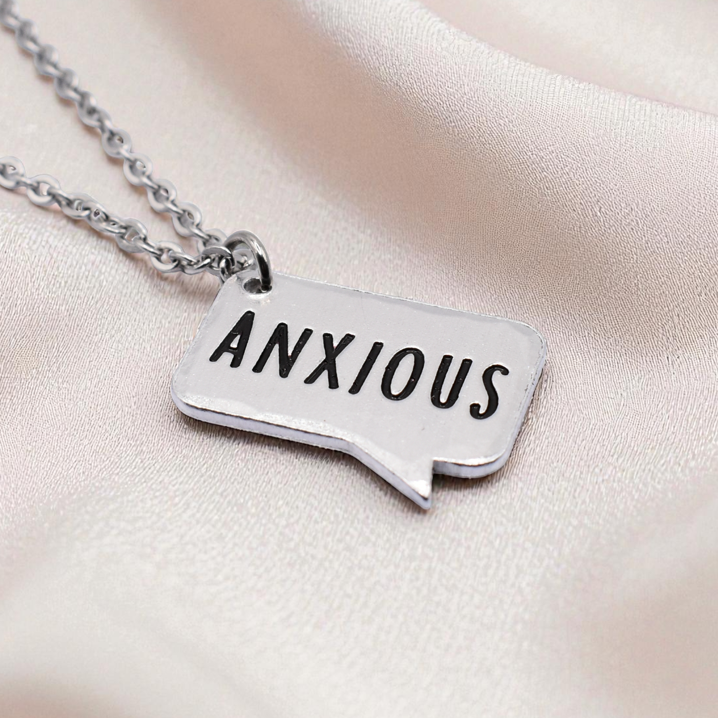 Silver necklace with a pendant that reads 'ANXIOUS' on a soft pink background