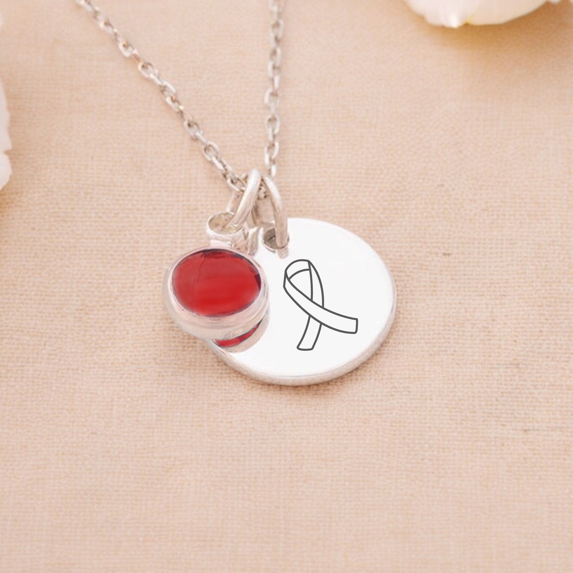 An Awareness Ribbon Necklace with a red birthstone charm placed on a chain, displayed with white flowers in the background.