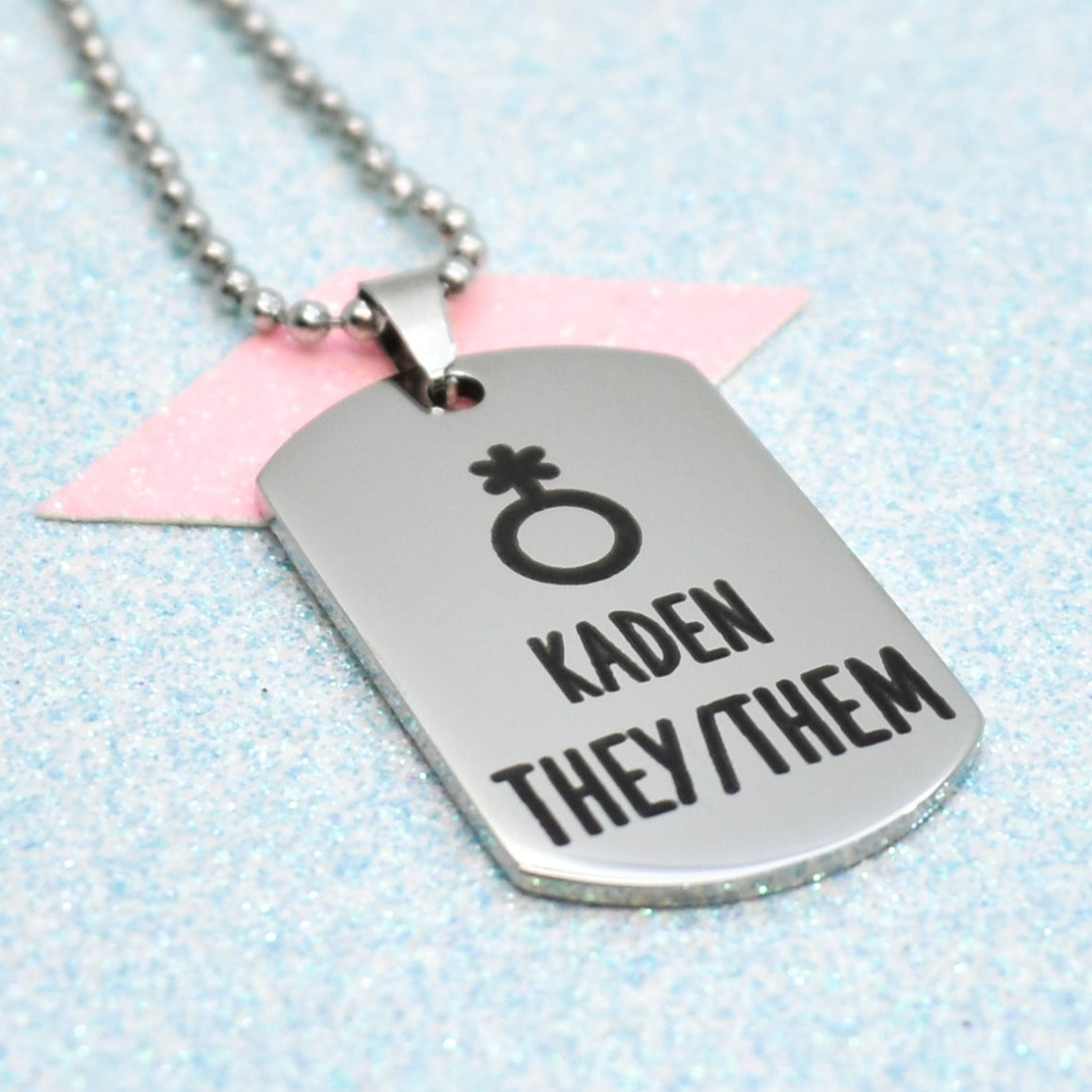 A silver necklace with a non-binary symbol and the text 'KadeN they/them' on it, displayed against a glittery blue background.