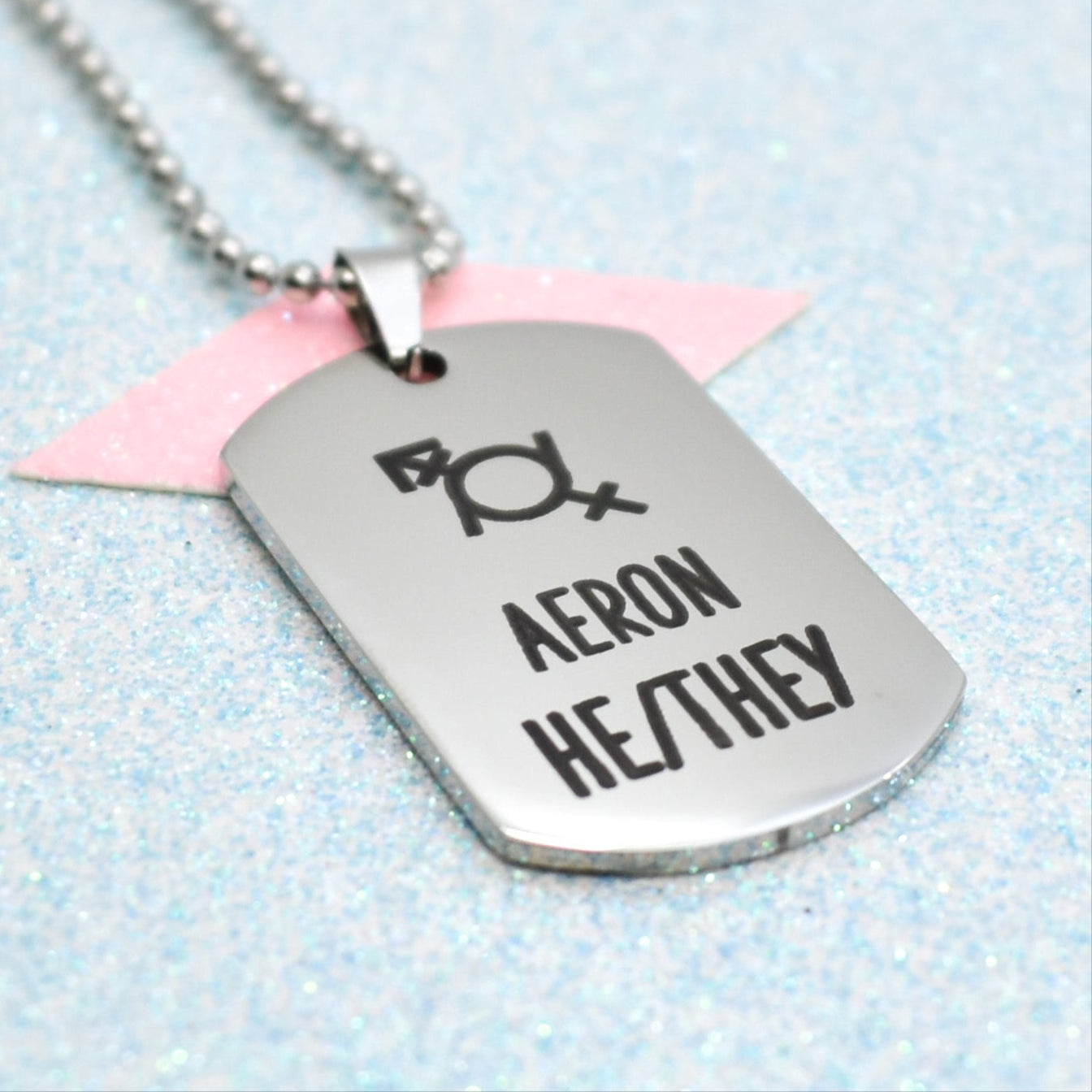 A silver necklace with a ball chain, featuring a stylized gender fluid gender symbol and the name 'AERON' in bold black letters.