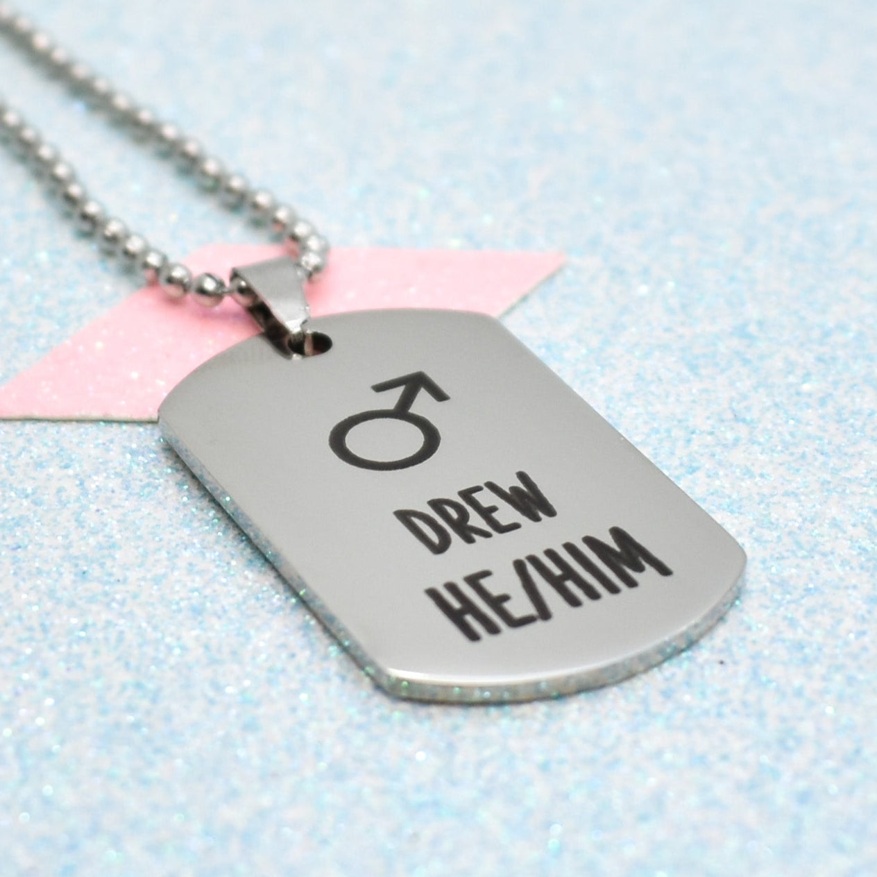 A silver necklace with a ball chain, featuring a stylized male gender symbol and the name 'Drew' in bold black letters.