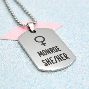 A silver necklace with a ball chain, featuring a stylized female gender symbol and the name 'Monroe' in bold black letters.