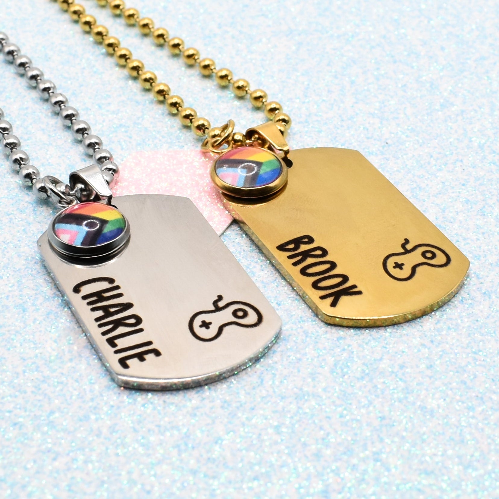 Two dog tag necklaces with engraved names and a pride flag charm on a light blue textured background