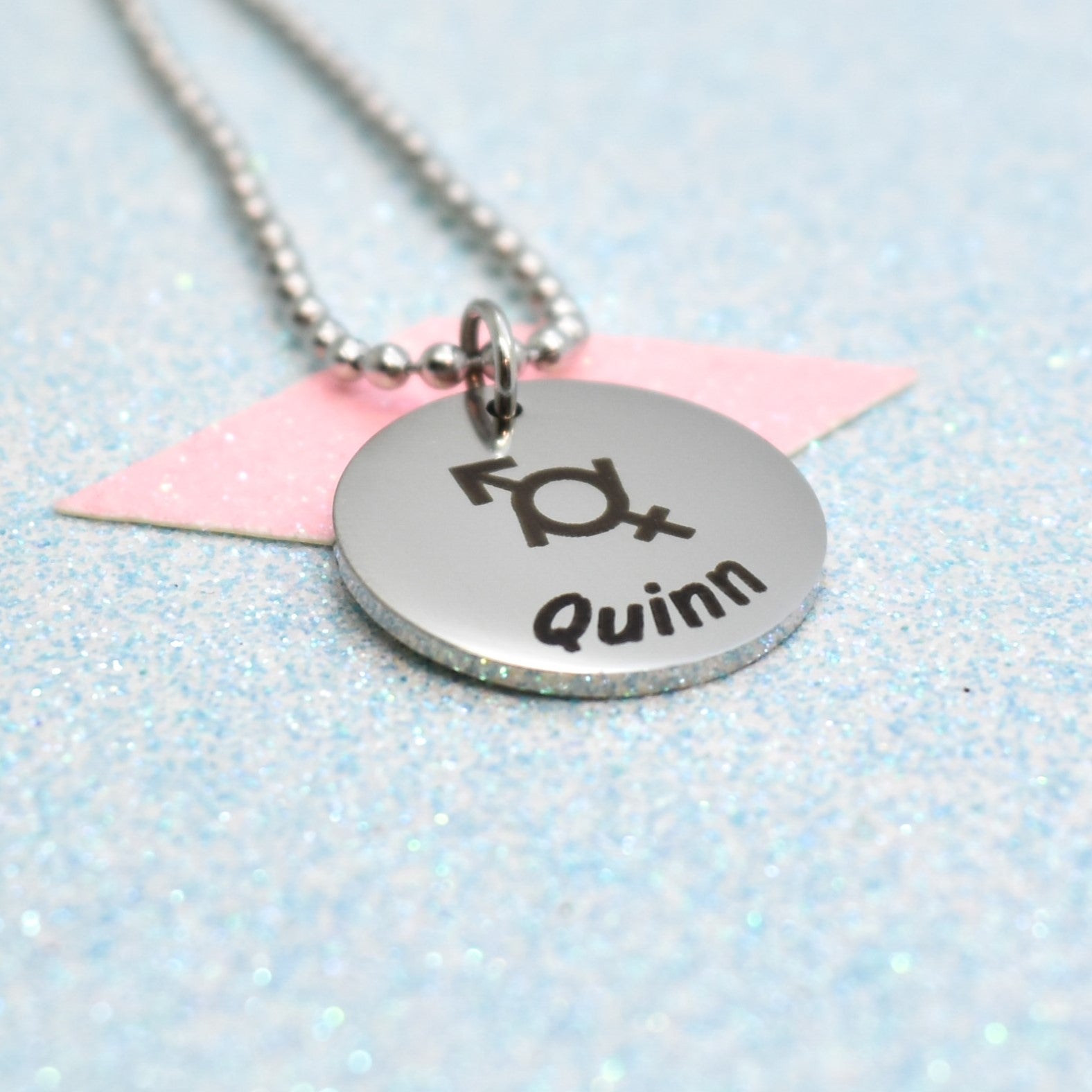A gender fluid necklace with a silver emblem and the name 'Quinn' on it, attached to a pink tag.