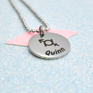 A gender fluid necklace with a silver emblem and the name 'Quinn' on it, attached to a pink tag.