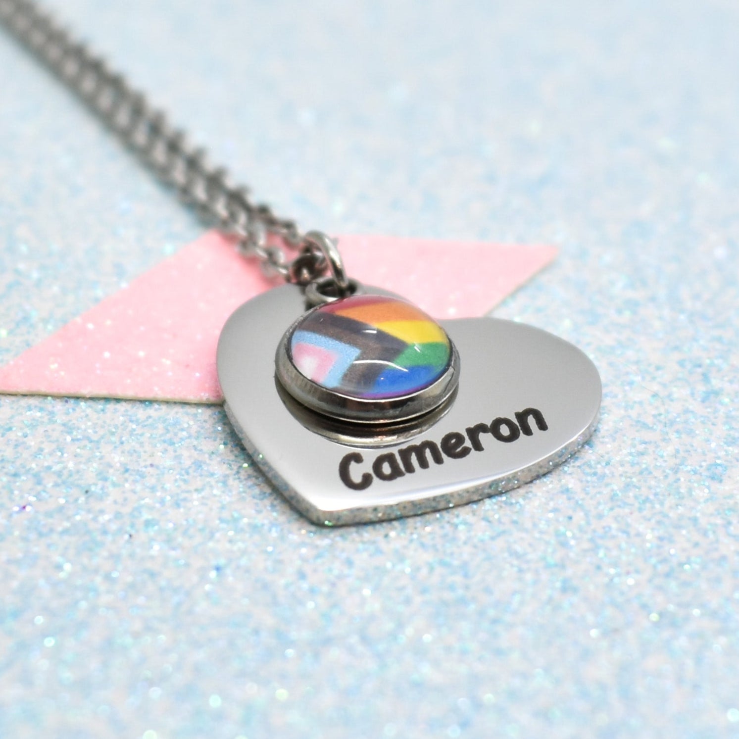 A heart-shaped pride necklace with a pride flag design and a personalised name name 'Cameron' on it, attached to a silver-coloured chain.