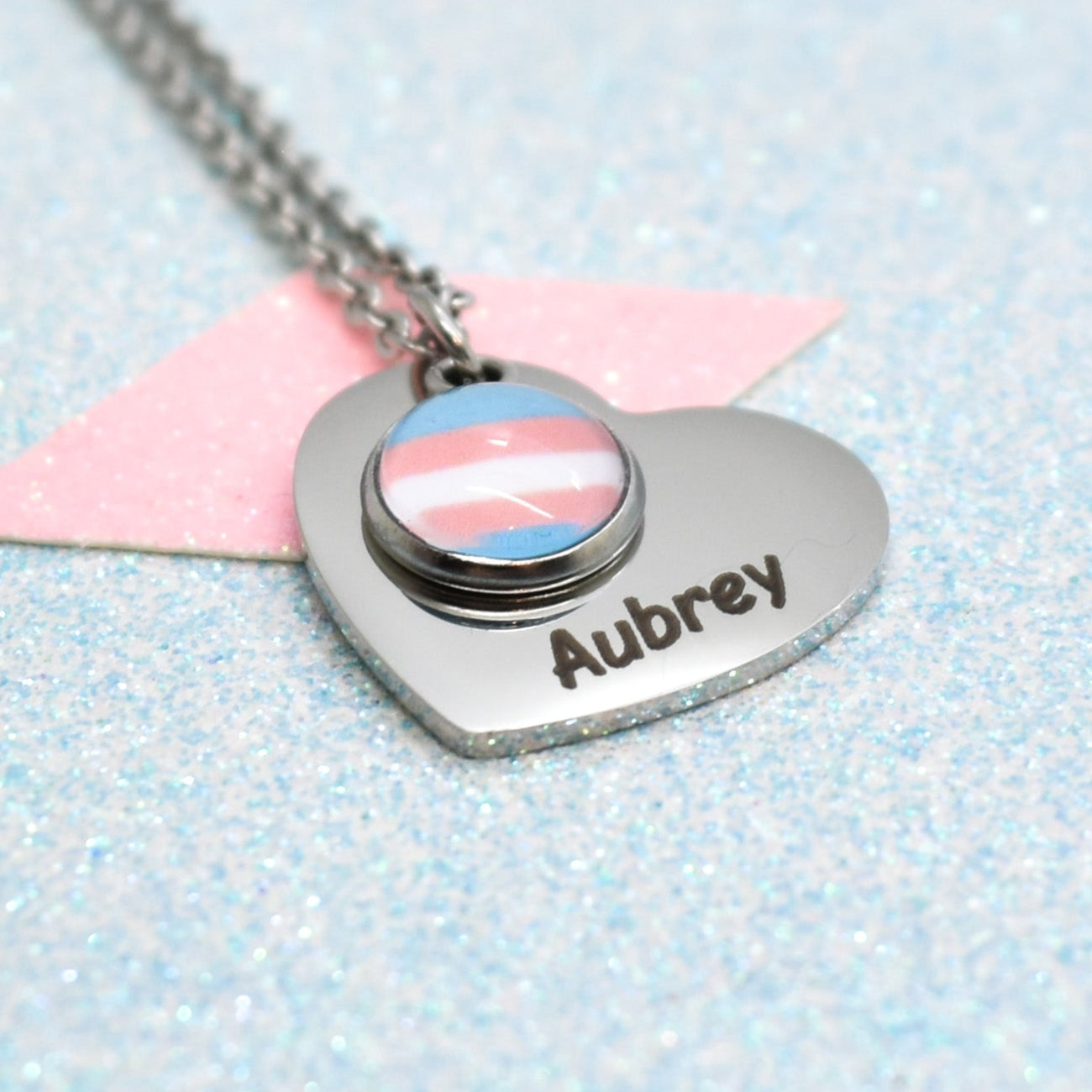 A heart-shaped transgender necklace with a pride flag design and the name 'Aubrey' on it, attached to a silver-coloured chain.