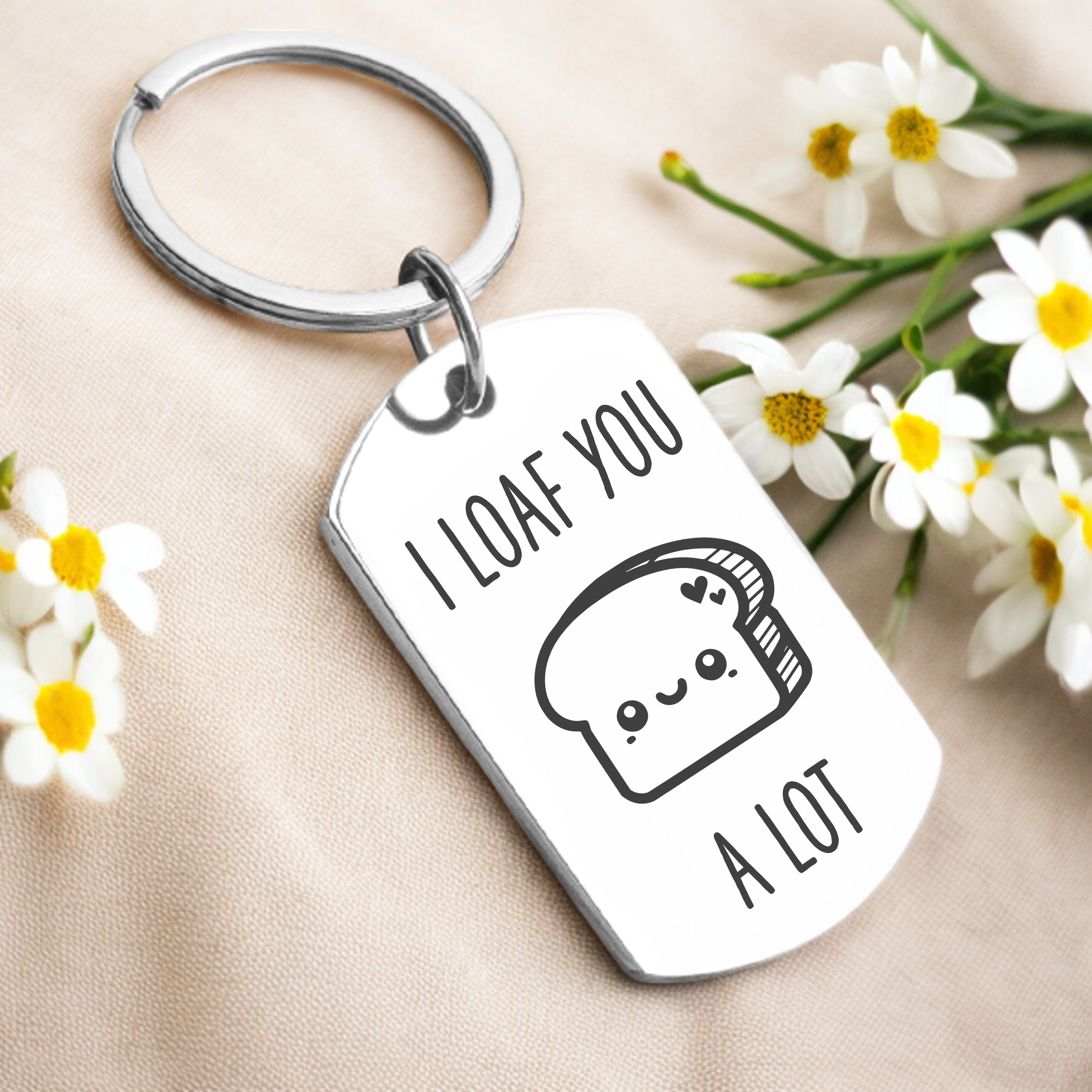 A personalized key ring with a silver or gold finish, featuring a playful design with a character and the phrase 'I loaf you a lot' on a rectangular tag, presented alongside daisies.