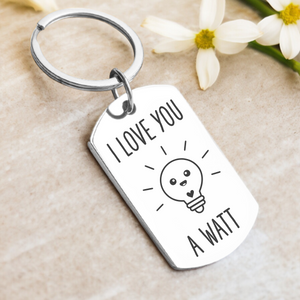 A key ring with a rectangular tag that has the phrase 'I LOVE YOU A WATT' printed on it, accompanied by a cartoon image of a light bulb. The tag appears to be made of silver and is displayed with a few petals at the bottom.
