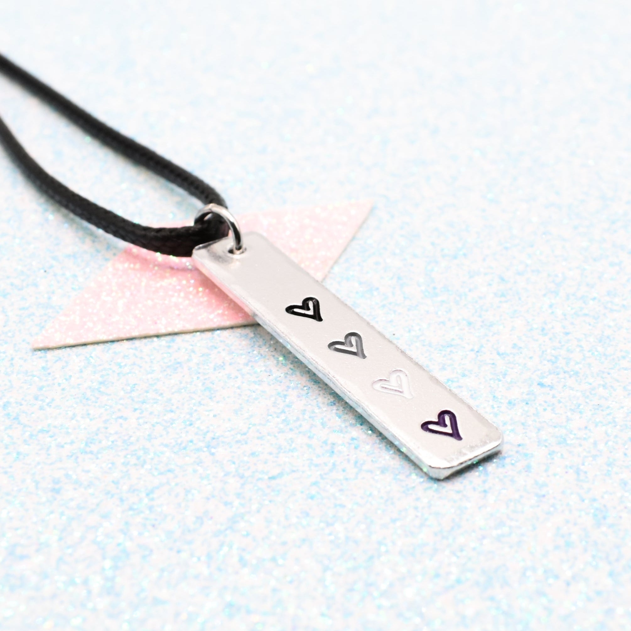 Necklace with a bar pendant on a faux leather cord featuring colourful hearts representing the asexual pride flag on a light blue background