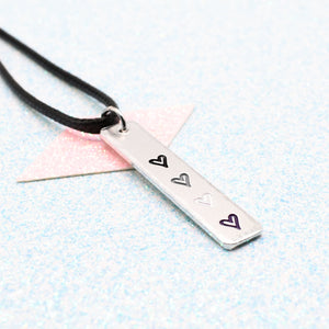 Necklace with a bar pendant on a faux leather cord featuring colourful hearts representing the asexual pride flag on a light blue background