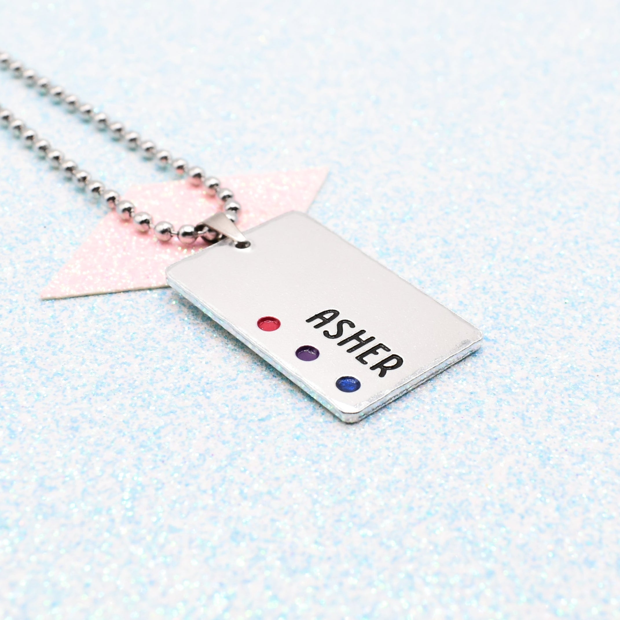 Customisable LGBTQ+ Pride Necklace featuring enamels of the Bisexual pride flag and a personalised name mounted on a ball chain displayed on a blue background