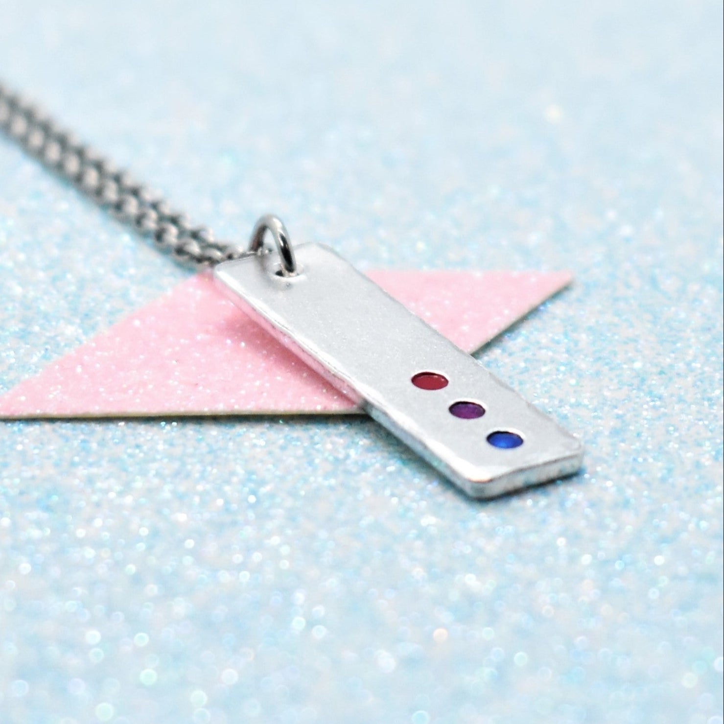 A minimalist pride necklace with a geometric shaped pendant featuring the bisexual flag colours, attached to a silver-coloured chain.