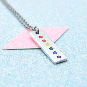 A minimalist pride necklace with a geometric shaped pendant featuring the rainbow flag colours, attached to a silver-coloured chain.