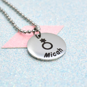 A non-binary necklace with a silver emblem featuring a gender symbol and the name 'Michah' on a chain.