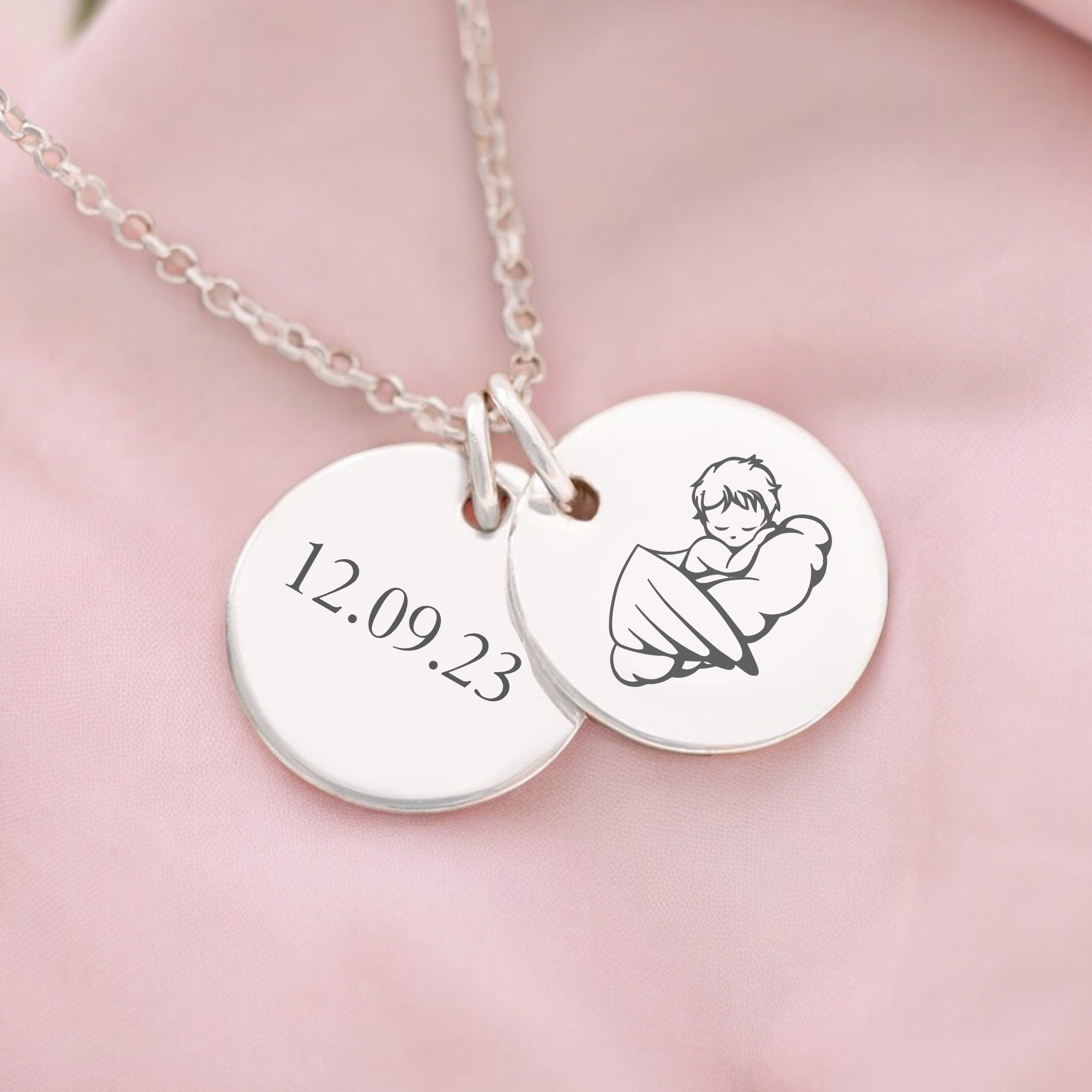 A personalised memorial necklace with two round discs, one engraved with a name or date and the other with an image of a baby. The necklace is displayed on a pink fabric.