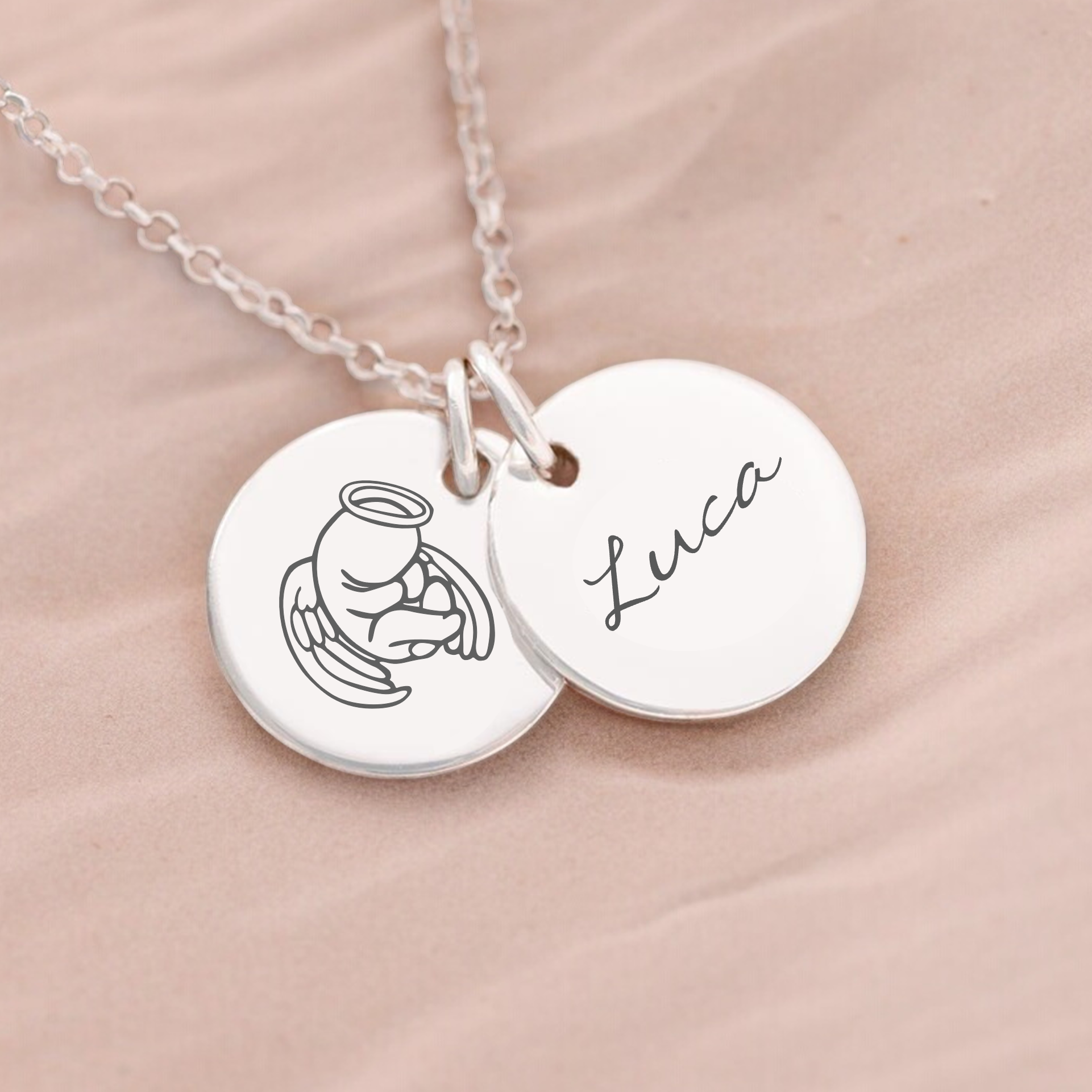 A silver memorial necklace with a personalized engraving of a name 'Luca' on a disc, featuring a small angel holding a heart charm, displayed on a beige background.