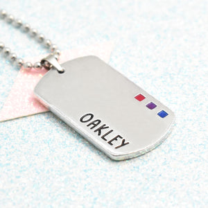 A silver-coloured necklace with a ball chain, featuring an enamelled bisexual flag badge and the name 'Oakley' written in bold letters.