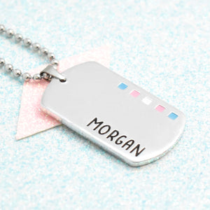 A silver-coloured necklace with an enamelled transgender flag design and the personalised name written on it.