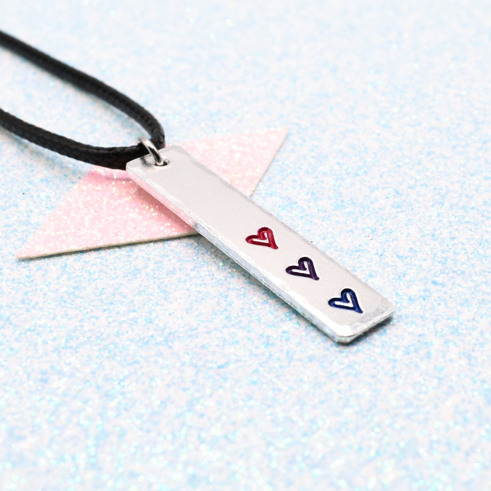 Necklace with a bar pendant on a faux leather cord featuring colourful hearts representing the bisexual pride flag on a light blue background