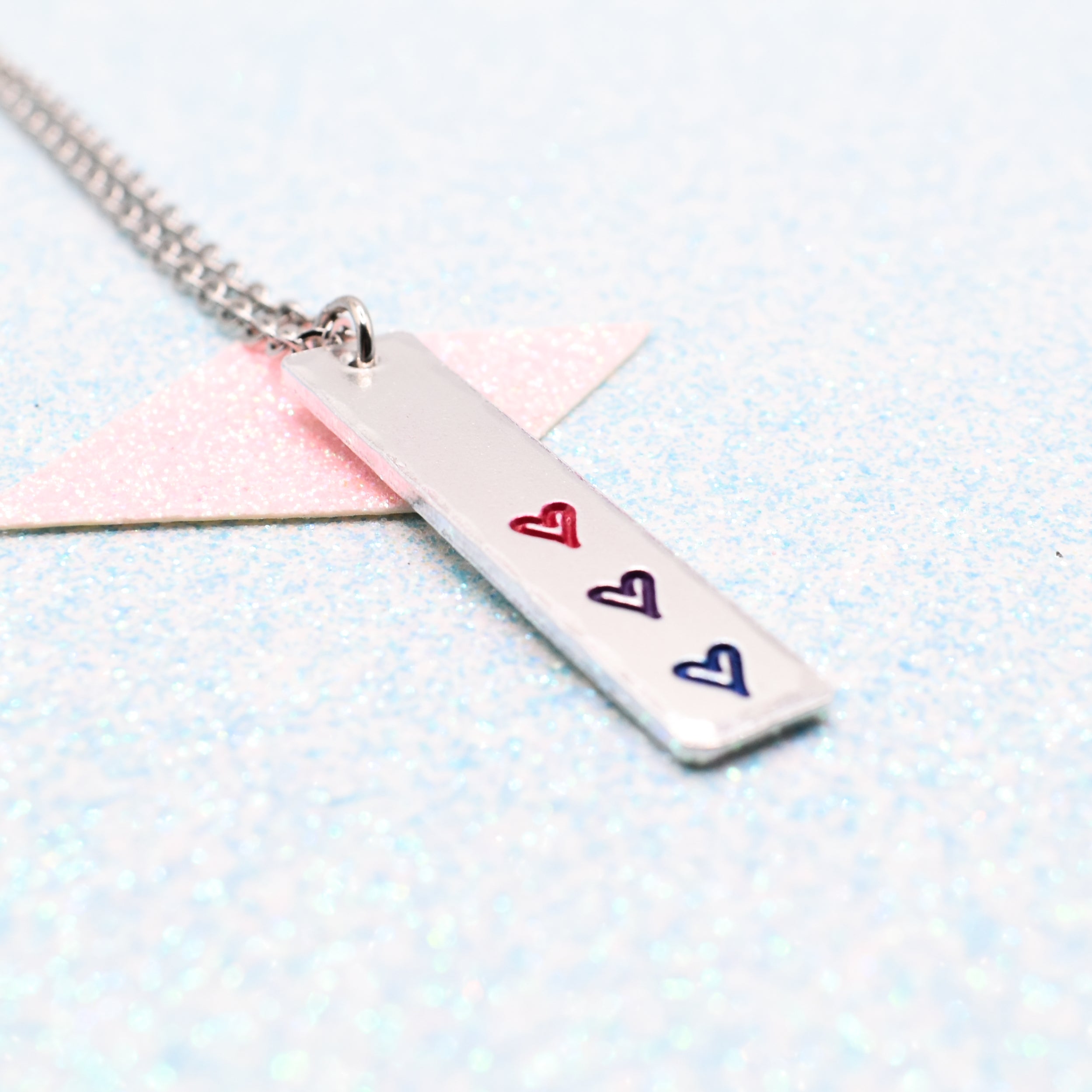 Necklace with a bar pendant on a ball chain featuring colourful hearts representing the bisexual pride flag on a light blue background